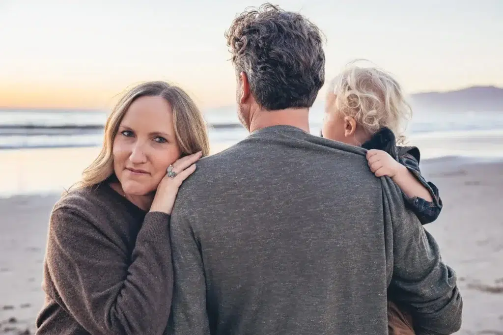 Heartwarming family moment on the beach at sunset at Santa Monica, California. A mother leans gently on her partner’s shoulder while their child, with curly blonde hair, clings to his sweater. The serene ocean waves and soft golden light create a warm and intimate atmosphere. 